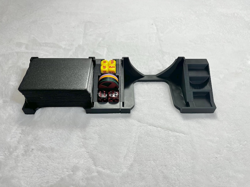May include: Black plastic tabletop game organizer with compartments for cards, dice, and other game accessories. The organizer has a rectangular card holder, a dice tray, and a curved section for additional items.