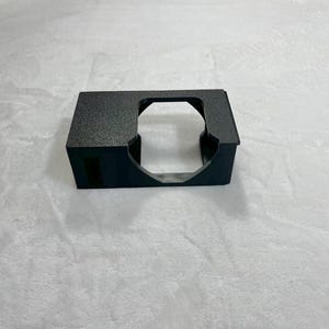 May include: A black, rectangular object with a textured surface and a large, circular opening. The object has a small, rectangular opening on one side. The object is sitting on a white surface.