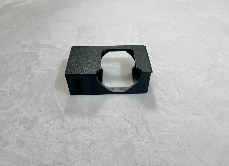 May include: A black, rectangular object with a textured surface and a large, circular opening. The object has a small, rectangular opening on one side. The object is sitting on a white surface.