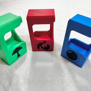 May include: Three colorful, angled card holders in green, red, and blue. The green holder has a tree silhouette, the red has a flame design, and the blue has a circular design. These are likely for trading card games.