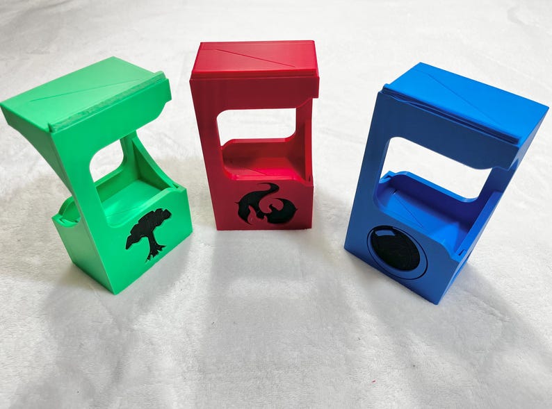 May include: Three colorful, angled card holders in green, red, and blue. The green holder has a tree silhouette, the red has a flame design, and the blue has a circular design. These are likely for trading card games.