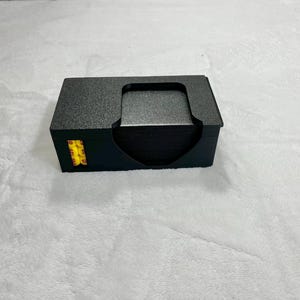 May include: A black card holder with a textured surface, holding a stack of black cards. The holder has a curved cutout for easy access to the cards. A small yellow and black sticker is visible on the side.