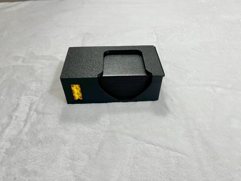 May include: A black card holder with a textured surface, holding a stack of black cards. The holder has a curved cutout for easy access to the cards. A small yellow and black sticker is visible on the side.