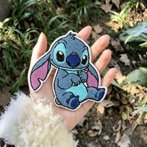 May include: An embroidered patch featuring the character Stitch with blue fur, a light blue chest, and pink inner ears. The design is outlined in white thread and depicts the character in a seated position with large black eyes and a dark blue nose.