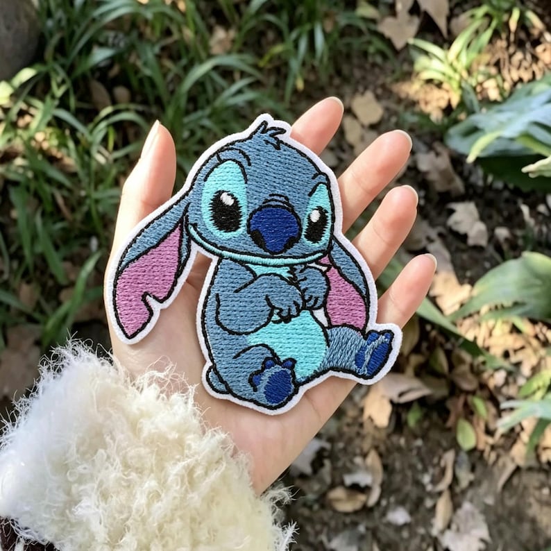 May include: An embroidered patch featuring the character Stitch with blue fur, a light blue chest, and pink inner ears. The design is outlined in white thread and depicts the character in a seated position with large black eyes and a dark blue nose.