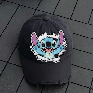 Puede incluir: Una gorra de béisbol negra con un parche bordado de un personaje alienígena azul con grandes orejas moradas y dientes afilados, que parece atravesar un efecto de papel blanco rasgado. La gorra tiene una visera desgastada y deshilachada.