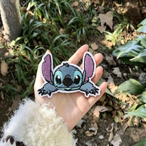 Op de afbeelding: Een geborduurde patch met het personage Stitch met blauwe vacht, grote paarse binnenoren en zwarte ogen. De patch heeft een witte rand en toont het personage in een liggende positie met de armen naar voren uitgestrekt, met gedetailleerd stikwerk.