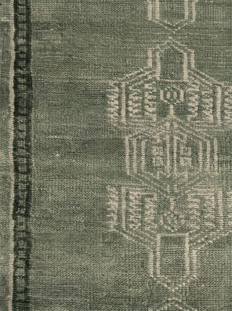 Op de afbeelding: Close-up van een groen en wit patroonkleed. Het kleed heeft een geometrisch ontwerp met een centraal patroon en een verticale rand aan de linkerkant. De textuur lijkt zacht en geweven.