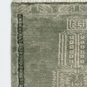 Op de afbeelding: Close-up van een saliegroen tapijt met een geometrisch patroon. Het tapijt heeft een getextureerd oppervlak en een rand met een donkerder groen ontwerp. De bovenrand heeft franjes. De woorden "Weave By Hand" staan in de linkerbovenhoek.