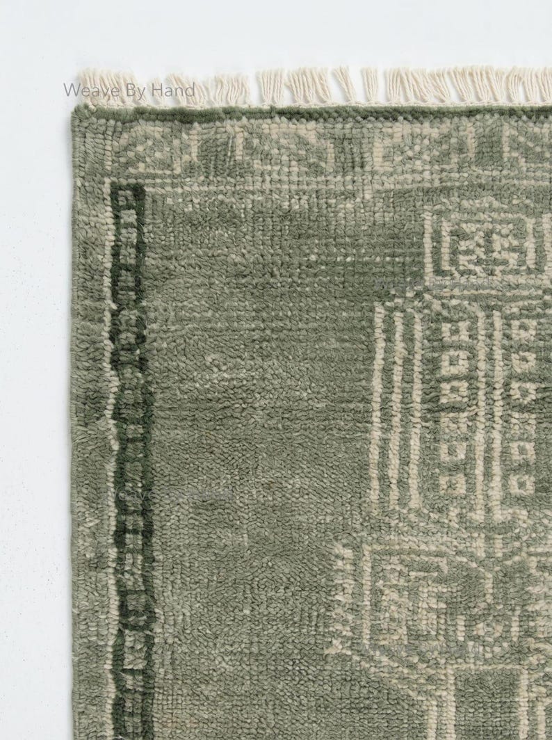 Op de afbeelding: Close-up van een saliegroen tapijt met een geometrisch patroon. Het tapijt heeft een getextureerd oppervlak en een rand met een donkerder groen ontwerp. De bovenrand heeft franjes. De woorden "Weave By Hand" staan in de linkerbovenhoek.