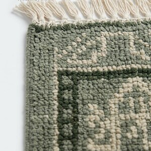 Op de afbeelding: Close-up van een groen en gebroken wit patroon tapijt met een franje. Het tapijt heeft een getextureerd uiterlijk en een geometrisch ontwerp. De tekst "Weave By Hand" is zichtbaar in de linkerbovenhoek.