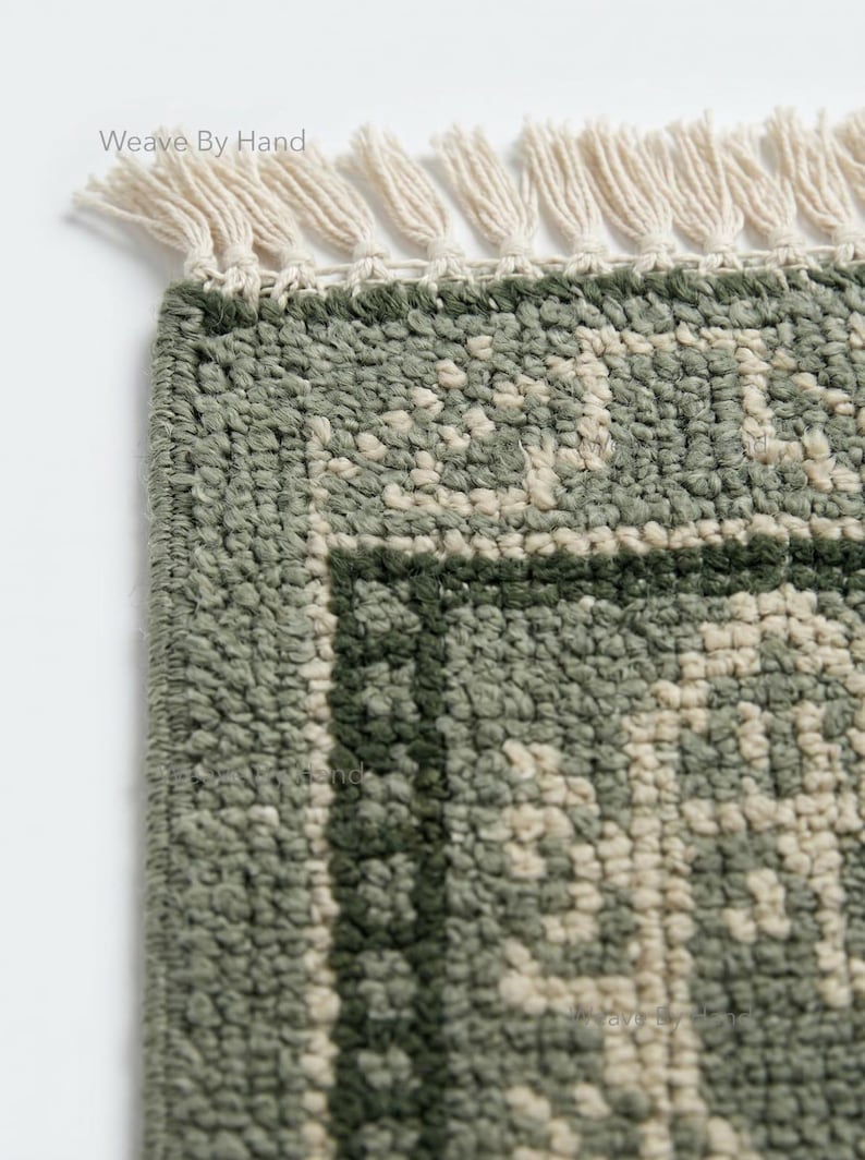 Op de afbeelding: Close-up van een groen en gebroken wit patroon tapijt met een franje. Het tapijt heeft een getextureerd uiterlijk en een geometrisch ontwerp. De tekst "Weave By Hand" is zichtbaar in de linkerbovenhoek.