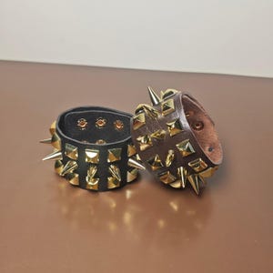 May include: Two wide wristbands featuring rows of metallic gold-toned spikes and pyramid studs. One band is black with a dark interior, and the other is brown with a lighter interior. Both wristbands include visible metal snap button closures.