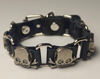 May include: Black leather bracelet with silver-colored skull and crossbones accents. The bracelet features a series of skull charms connected by metal links and a buckle closure. The design has a gothic or punk rock aesthetic.