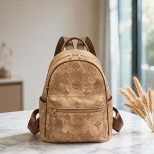 May include: A tan backpack featuring a repeating embossed floral pattern across the exterior. The bag includes a front zippered pocket with a gold-toned metal pull, two side slip pockets, and adjustable brown shoulder straps with gold hardware.