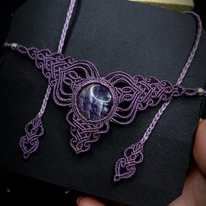 Può includere: Una collana in macramè viola fatta a mano con una pietra rotonda lucida centrale. L'intricato design in macramè include elementi decorativi ed estensioni del cordoncino. La collana è esposta su una superficie nera.