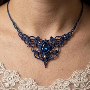 May include: A navy blue macrame necklace with a large, teardrop-shaped, dark blue gemstone at the center. The necklace features intricate knotwork and small silver beads. The necklace is worn around the neck.