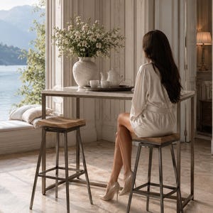 May include: A contemporary bar table with a marble surface and metal frame, with two wooden and metal stools. A white vase with white flowers is on the table, alongside a tea set. The room is bright, with a view of a lake in the background.