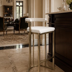 May include: A modern bar stool with a white seat and backrest. The frame is made of brushed metal. The stool has a curved base and a rectangular footrest. The stool is in a well-lit room with a marble floor.