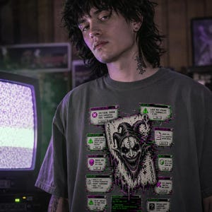May include: A gray t-shirt featuring a graphic of a Joker playing card with a sinister clown face and glitching computer windows. The shirt has the word "JOKER" vertically on the left side. The design incorporates green, purple, and white elements.
