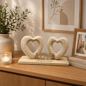Könnte beinhalten: Zwei herzförmige Holzskulpturen mit der Aufschrift 'LOVE' und den Namen 'Adrian & Elena' auf einem Holzsockel. Die Herzen sind hell, und die Szene umfasst eine Kerze, eine Vase mit Grün und gerahmte Kunstwerke.