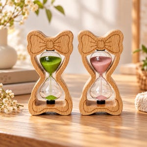 May include: Two hourglasses with light-colored wooden frames shaped like bows. The left hourglass contains vibrant green sand, while the right hourglass contains soft pink sand. Both glass bulbs are encased in smooth, carved wood structures.