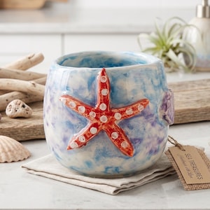 May include: A ceramic tumbler with a red starfish design. The tumbler has a blue and white watercolor effect with a purple accent. The tag reads "Sea Treasures Handmade with Love".