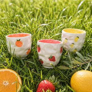Könnte beinhalten: Drei weiße Keramikbecher mit Fruchtdesign auf grünem Gras. Einer hat Orangen, ein anderer Erdbeeren und der dritte Zitronen. Die Innenräume sind orange, rot und gelb. Eine aufgeschnittene Orange, eine Erdbeere und eine Zitrone sind ebenfalls zu sehen.