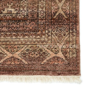 Pode incluir: Vista de perto de uma secção de um tapete vintage com um padrão geométrico detalhado. O tapete apresenta uma rica paleta de cores de bordô, bege e castanho, com uma franja creme na borda inferior. O design inclui motivos repetidos de diamantes e flores.