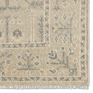 May include: Close-up of a beige and grey patterned rug. The rug has a textured surface and a fringe along the bottom edge. The design features repeating shapes and floral motifs in a muted colour palette.