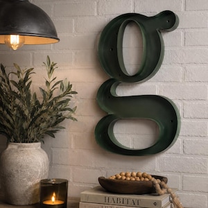 May include: A dark green metal wall-mounted letter g with a hollow interior. The letter features a smooth finish and mounting holes along its frame, designed for vertical display on a wall surface as a decorative piece of signage or wall art.
