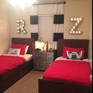 May include: Two twin beds with red bedding and wooden headboards. The beds are in a room with black and white striped curtains and a metal locker style nightstand. The room is decorated with lighted letter signs that spell "RZ".
