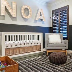 May include: A white crib with a brown and white patterned sheet and a white crib skirt. The crib is in a nursery with a gray and white striped rug, a gray rocking chair with a white pillow, and a brown ottoman. The wall is painted black and white with the name "NOAH" spelled out in white lights.