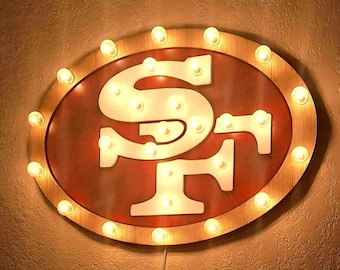 May include: An oval-shaped wall sign featuring the San Francisco 49ers logo in white against a dark red background. The sign is framed by a metallic border illuminated by a series of small, warm-toned light bulbs spaced evenly around the perimeter.