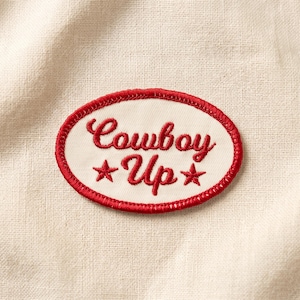 May include: An oval-shaped embroidered patch featuring a cream-colored background with a red stitched border. The text "Cowboy Up" is embroidered in red cursive script, flanked by two small red embroidered stars on either side of the word Up.