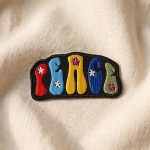 May include: A colorful embroidered patch with the word "PEACE" in a playful font. The letters are in red, blue, yellow, blue, and green, each with a small flower accent. The patch has a black border and is set against a cream-colored fabric.