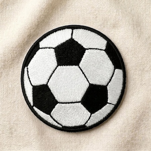 May include: A circular embroidered patch featuring a classic soccer ball design with alternating black pentagons and white hexagons. The stitching is dense and uniform, creating a textured surface with a distinct black border around the edge.
