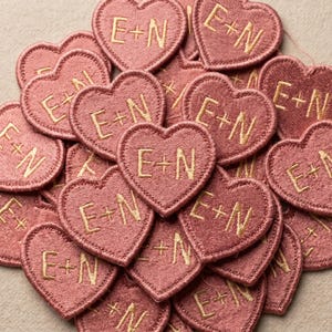 May include: A collection of heart-shaped patches in a dusty rose color. Each patch has the embroidered letters "E+N" in a gold color. The patches are closely arranged, creating a textured surface.