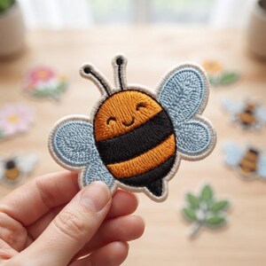 May include: An embroidered patch shaped like a cartoon bee with a round body featuring horizontal orange and black stripes, two light blue wings, and two antennae. The bee has a simple stitched face with closed eyes and a small curved smile.