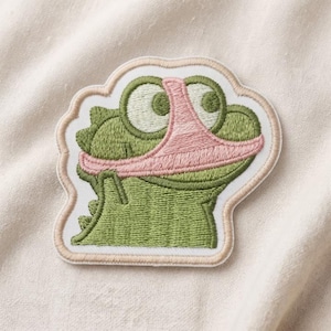 May include: An embroidered patch featuring a green cartoon creature with large eyes and a pink thong worn over its face like a mask. The patch has a light beige border and is shaped to follow the outline of the character's head and the garment.