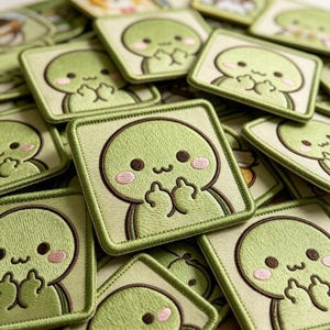 May include: A square embroidered patch featuring a light green cartoon character with a round head, dark brown eyes, a small mouth, and pink cheeks. The character is holding up both middle fingers. The patch has a matching green stitched border.