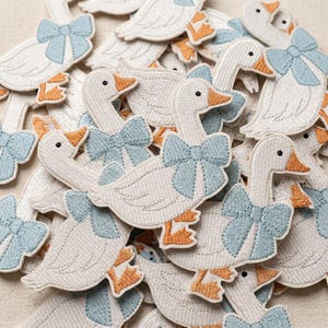 May include: A collection of white goose patches with light blue bows and orange feet. The patches are embroidered with detailed stitching, creating a textured appearance. The geese are in various poses, creating a charming and whimsical design.