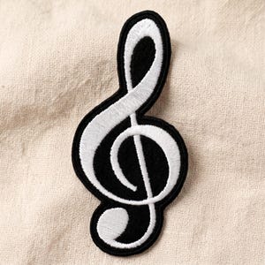 May include: A black and white embroidered patch in the shape of a treble clef musical symbol. The design features a solid black background with a thick, raised white thread outline and interior detailing that follows the classic musical notation form.