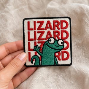 May include: Square patch with a black border featuring the word "LIZARD" in red, repeated three times. A green cartoon lizard with big eyes and a waving hand is in the lower right corner. The patch is on a beige fabric.