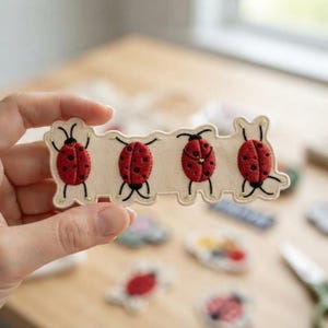 May include: An elongated, cream-colored fabric patch featuring four embroidered red ladybugs with black spots and black legs. The patch has a scalloped, irregular edge that follows the outline of the ladybug figures, creating a continuous strip.
