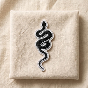 May include: Black and silver embroidered snake patch. The snake design is in a vertical, coiled position with a silver border. The patch is on a beige fabric background.
