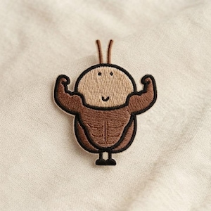 May include: An embroidered patch featuring a cartoon character with a tan head, brown muscular arms, and a brown torso. The character has two antennae, a simple smiling face, and is depicted in a flexing pose with a thick black outline.