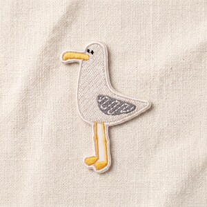 May include: An embroidered patch featuring a white seagull with a yellow beak and feet, and grey wing detailing. The bird is depicted in profile facing left, with a simple black eye, stitched onto a textured, off-white fabric background.