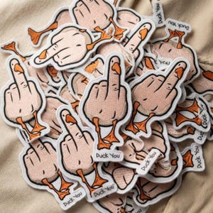 May include: A collection of embroidered patches featuring a light-skinned hand making a middle finger gesture with an orange duck beak and orange webbed feet attached. Each patch includes the text Duck You printed in black at the bottom.