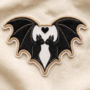 May include: An embroidered patch featuring two black bats facing each other with wings spread wide to form a heart shape. A small black heart is positioned between the bats' heads. The design is outlined in gold and white thread.